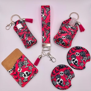 Puede incluir: Un conjunto de accesorios con un diseño de calaveras y rosas en rosa, negro y blanco. Incluye una correa de muñeca, llaveros con borlas, un tarjetero y posavasos para coche. Tema gótico.