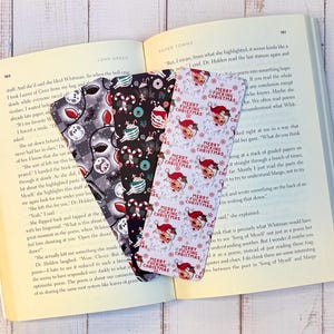 Op de afbeelding: Drie decoratieve bladwijzers met unieke patronen worden in een open boek getoond. Eén bladwijzer heeft een Halloween-thema, een andere een kerstontwerp en de derde een kerstmanontwerp met de tekst "MERRY FUCKING CHRISTMAS".