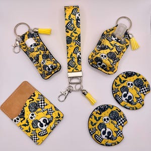 Op de afbeelding: Een set accessoires met een gele achtergrond en een zwart-wit patroon van schedels, rozen, vleermuizen, messen en geblokte harten. Bevat een polsband-sleutelhanger, een lippenbalsemhouder, een handgelhouder, een kaarthouder en twee ronde auto-onderzetters, allemaal met bijpassende gele kwastjes.