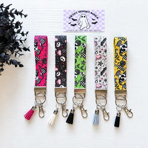 May include: Five wristlet keychains featuring patterned fabric straps in pink, black, green, white, and yellow. Each strap has a silver-tone metal clasp, a split ring, a lobster claw hook, and a small hanging tassel in matching colors.