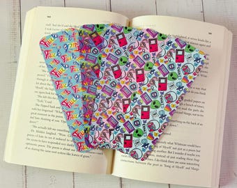 Pode incluir: Um conjunto de marcadores coloridos com vários padrões, incluindo a palavra "Fuck" em cores do arco-íris e um design com consolas de videojogos retro, alienígenas e outros elementos nostálgicos, colocados num livro aberto.