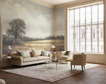 Op de afbeelding: Een woonkamer met een grote muurschildering die een landschap met een boom en een veld afbeeldt. De muurschildering bevindt zich op de muur achter een beige bank en twee fauteuils. De kamer heeft een groot raam met uitzicht op een gebouw. Er ligt een tapijt op de vloer en voor de bank staat een salontafel.