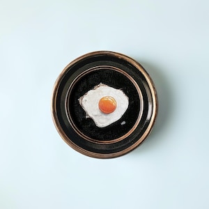 May include: A framed painting of a fried egg on a black background. The egg yolk is a bright orange color and the white is a creamy white. The frame is a dark brown wood with a gold trim.