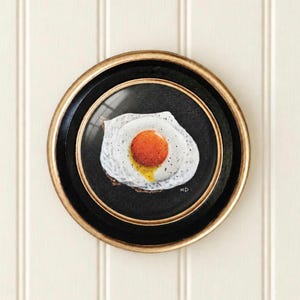 May include: A circular framed artwork featuring a realistic painting of a fried egg with a bright orange yolk and white edges on a solid black background. The frame is gold with a black inner border. The initials HD are visible in white.