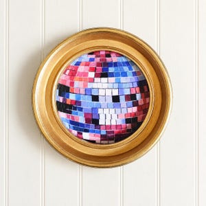 May include: A circular gold frame containing a round artwork depicting a disco ball. The disco ball is composed of small, square tiles in shades of pink, blue, purple, white, and black, creating a reflective, multi-faceted appearance.