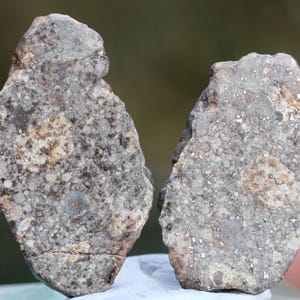 Pode incluir: Duas rochas de forma irregular, de cor cinza, com uma superfície rugosa e texturizada. As rochas têm uma mistura de cores, incluindo castanho e branco, e parecem ser fragmentos de uma pedra maior. O fundo está desfocado.
