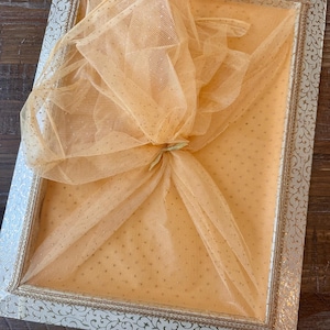 Puede incluir: Una caja de regalo rectangular con un marco de color crema y un intrincado patrón de filigrana dorada. El interior está forrado con una tela suave de color melocotón, cubierta por una red de tul transparente atada en el centro.