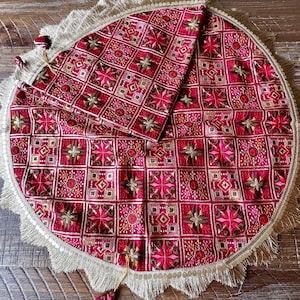 May include: A circular fuchsia fabric mat featuring a grid pattern of embroidered geometric shapes and floral motifs in shades of pink, red, and gold. The edge is finished with a wide beige fringe and a decorative trim with small gold sequins.
