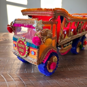 May include: A decorative, colorful toy truck with intricate detailing. The truck features a vibrant mix of orange, pink, yellow, and blue, adorned with gold trim, pom-poms, and a star-shaped embellishment. The front displays the text "Happy Lohri".