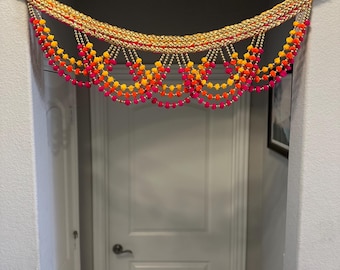 Puede incluir: Un colgador de puerta colorido con pompones rosas, naranjas y amarillos y detalles dorados. El colgador está hecho de tela y cuentas y está colgado en un marco de puerta blanco.
