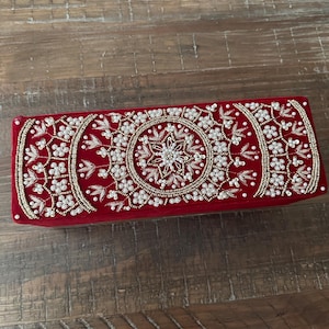May include: A rectangular, red velvet clutch with intricate beadwork. The design features a central floral motif surrounded by a circular pattern and embellished with white beads and gold accents. The clutch is a decorative accessory.