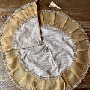 May include: A circular off-white fabric cover with a textured geometric pattern, featuring a wide, pleated gold-toned sheer border trimmed with metallic gold lace. Two silver-toned flower-shaped embellishments with red centers are attached.