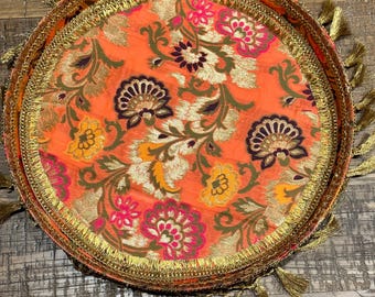 May include: A round, orange tray with a floral pattern and gold trim. The tray has a fringe border with gold tassels.