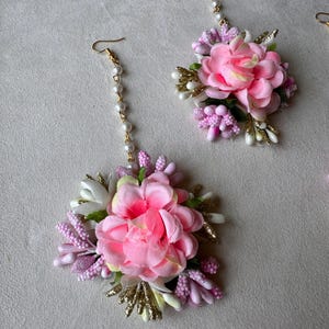 May include: Floral earrings featuring large pink fabric flowers surrounded by smaller pink and white accents, with gold-colored accents and a pearl chain. The earrings have a hook closure.