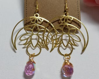Puede incluir: Pendientes dorados con un diseño floral y de media luna. Cada pendiente presenta un cristal rosa iridiscente colgante. Los pendientes se exhiben en una tarjeta marrón que dice "Handmade with love."