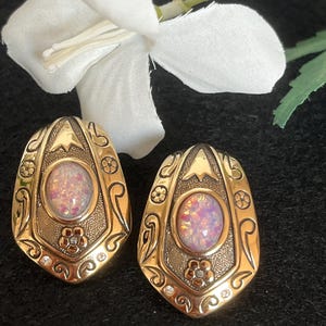 Könnte beinhalten: Ein Paar goldfarbene Ohrringe in Schildform mit einem ovalen, irisierenden künstlichen Opal in der Mitte. Die Metalloberfläche zeigt gravierte Schnörkel, kleine Blumenmotive und winzige klare Steine, die an der Basis jedes Ohrrings eingelassen sind.