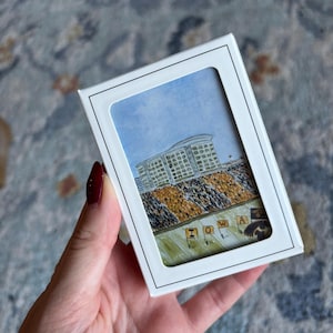 May include: A framed watercolor painting of a stadium with the word "IOWA" visible. The painting is in a white frame with a black border. The stadium has a blue sky background and is filled with people wearing gold and black.