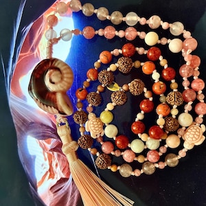 Könnte beinhalten: Eine mehrsträngige Mala-Halskette mit einem Quaste. Die Halskette besteht aus verschiedenen Perlen, darunter rosa, rote, braune und grüne. Die Quaste ist aus hellbraunem Material.