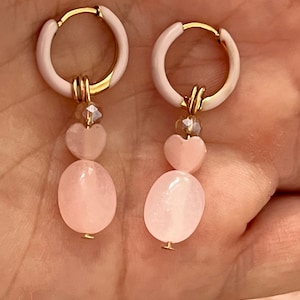 Op de afbeelding: Een paar gouden creolen met roze emaille en een roze hart- en ovale hanger die aan elke creool hangt.