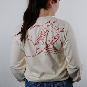 Può includere: Una maglietta a maniche lunghe color crema con un'illustrazione rossa di una manta gigante sul retro. L'illustrazione include il testo "Giant manta ray Mobula birostris".