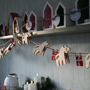 Op de afbeelding: Een decoratieve slinger van houten Dala-paarden met kleine lampjes, geregen aan een dunne draad. De paarden zijn lichtgekleurd hout met geëtste details. Kleine rood-witte cadeau-vormige ornamenten staan ook op de slinger. Op de achtergrond staan kleine huisvormige kaarshouders.