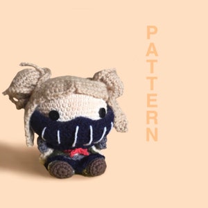 May include: A crocheted plush toy of a character with blonde hair, a black mask, and a red bow tie. The toy is sitting on a light brown surface. PATTERN is written vertically in orange letters on the right side of the image.
