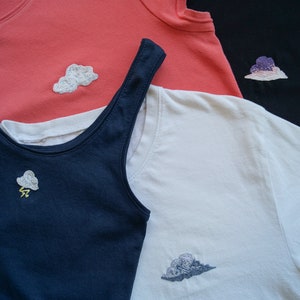 Può includere: Tre canottiere diverse con disegni di nuvole ricamate. Le canottiere sono blu navy, bianche e rosa corallo. La canottiera blu navy ha un fulmine sotto la nuvola.