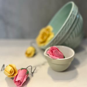 May include: A close-up of three pastel-colored fabric roses, two yellow and one pink, with one rose in a small white ceramic bowl. The bowl is sitting on a white surface. A larger, light green ceramic bowl is in the background.