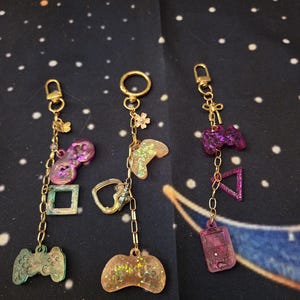 May include: Three gold-toned metal keychains featuring resin charms in shapes of video game controllers, squares, triangles, and rectangles. The charms are translucent with glitter and purple, green, and tan hues, attached to gold chains with lobster clasps.