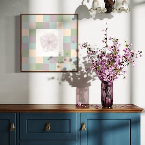 Könnte beinhalten: Ein gerahmtes Kunstwerk mit floralem Design hängt über einem blauen Schrank. Eine Vase mit rosa Blumen steht auf dem Schrank. Das Kunstwerk zeigt eine zentrale Blumenskizze, umgeben von einem Raster aus Pastellquadraten. Eine dekorative Wandleuchte ist darüber angebracht.
