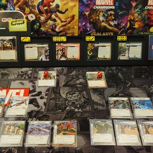 May include: A Marvel Champions game in progress with cards laid out on a black and white game mat. The game board is in the center of the image with dice and character cards. There are two players with their decks of cards in boxes on either side of the game board. The game board has the words "Marvel Champions" printed on it.