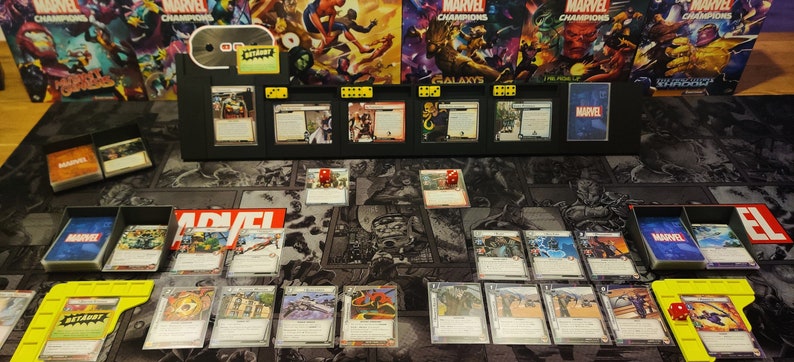 May include: A Marvel Champions game in progress with cards laid out on a black and white game mat. The game board is in the center of the image with dice and character cards. There are two players with their decks of cards in boxes on either side of the game board. The game board has the words "Marvel Champions" printed on it.
