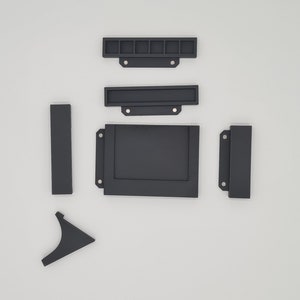 May include: A set of six black plastic pieces for a craft project. The pieces include a rectangular frame, a smaller rectangular frame, a long thin rectangle, a short thin rectangle, and two L-shaped pieces.
