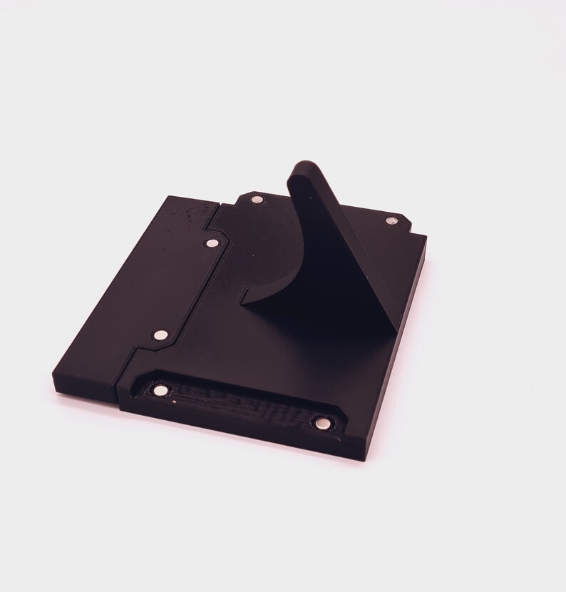 May include: A black plastic stand with a raised, angled platform. The stand has several small, round, silver-colored dots on the surface.