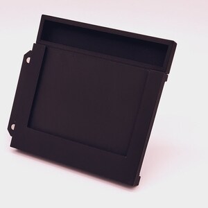 May include: A black plastic case with a slot for a game cartridge. The case has a hinged lid and a small compartment on the top.