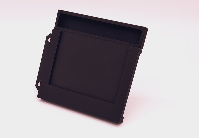 May include: A black plastic case with a slot for a game cartridge. The case has a hinged lid and a small compartment on the top.