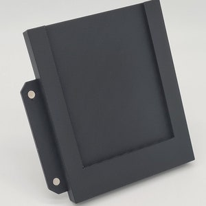 May include: A black plastic picture frame with a magnetic closure on the side. The frame is designed to stand upright on a flat surface.