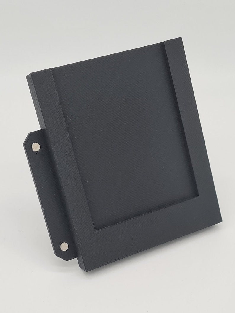 May include: A black plastic picture frame with a magnetic closure on the side. The frame is designed to stand upright on a flat surface.