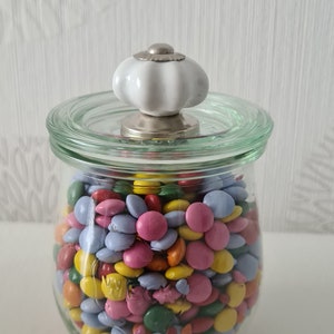 Könnte beinhalten: Ein klares Glas mit bunten Süßigkeiten gefüllt. Das Glas hat einen Glasdeckel mit einem weißen, abgerundeten Knauf. Die Süßigkeiten sind in verschiedenen Farben, darunter Rosa, Blau, Gelb und Orange.