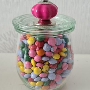 Könnte beinhalten: Ein Glasgefäß mit einem rosafarbenen Knopfdeckel, gefüllt mit bunten, zuckerüberzogenen Schokoladenstückchen.