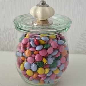 Könnte beinhalten: Ein Glasgefäß mit einem weißen Keramikdeckel mit Knopf, gefüllt mit bunten Süßigkeiten. Die Süßigkeiten sind ein Mix aus rosa, gelb, blau, grün und orange.