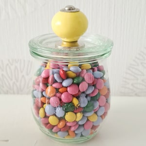 Könnte beinhalten: Ein Glasgefäß mit einem gelben Knopfdeckel gefüllt mit bunten Süßigkeiten. Die Süßigkeiten sind ein Mix aus Rosa, Blau, Grün, Gelb und Orange.