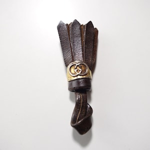 Puede incluir: Un borla de cuero marrón con un dije de oro y plata con el logotipo G entrelazado. La borla está anudada en la parte inferior.