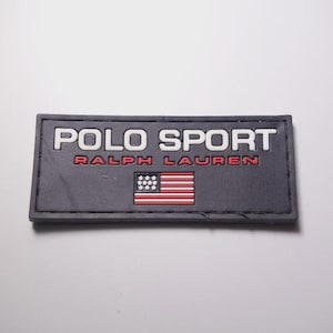 May include: A rectangular black rubber patch featuring the white embossed text POLO SPORT and the red text RALPH LAUREN. Below the text is a small, raised graphic of the American flag with white stars and red and white horizontal stripes.