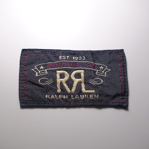 Puede incluir: Un parche de mezclilla azul con costuras blancas y bordados rojos. El parche presenta el texto "EST. 1993" y "DOUBLE RL" con una estrella a cada lado del texto. El texto "RALPH LAUREN" está debajo del logotipo.