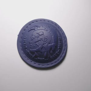 May include: A circular purple patch featuring an embossed profile of a mohawked figure. The text "ONLY THE BRAVE" and "DIESEL" is molded into the border surrounding the central design. The patch has visible stitching along the outer edge.