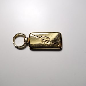 May include: A gold-colored key chain with a rectangular tag featuring the Gucci logo. The tag has a diagonal line across it.