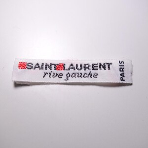 Puede incluir: Etiqueta tejida blanca con texto negro que dice "SAINT LAURENT rive gauche PARIS". Las palabras "SAINT" y "LAURENT" están en rojo.