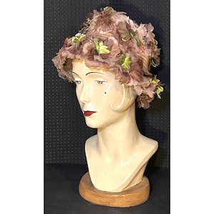 May include: A cloche hat covered in layered mauve fabric petals and small green leaves. The hat features a structured crown and a slightly downturned brim, with the floral elements densely arranged across the entire surface of the headwear.