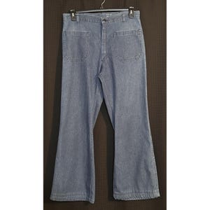 May include: Light blue denim jeans with a flared leg design. The jeans have two large back pockets and two front pockets. The jeans are hanging on a black hanger against a dark brown background.